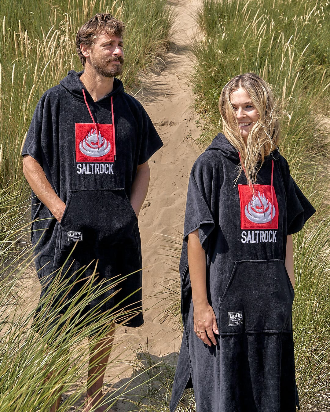 Two individuals wearing Saltrock Corp Changing Towel - Black hooded beach towels stand on a sandy path surrounded by tall grass.