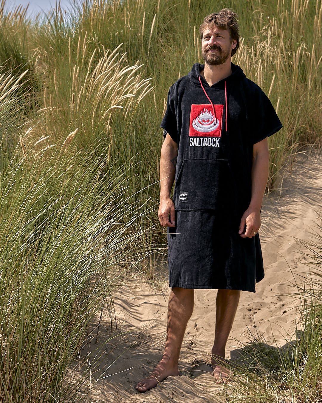 A man in a black Corp Changing Towel - Black made of absorbent, cotton towelling material with a Saltrock logo standing on a sandy beach with tall grasses in the background.