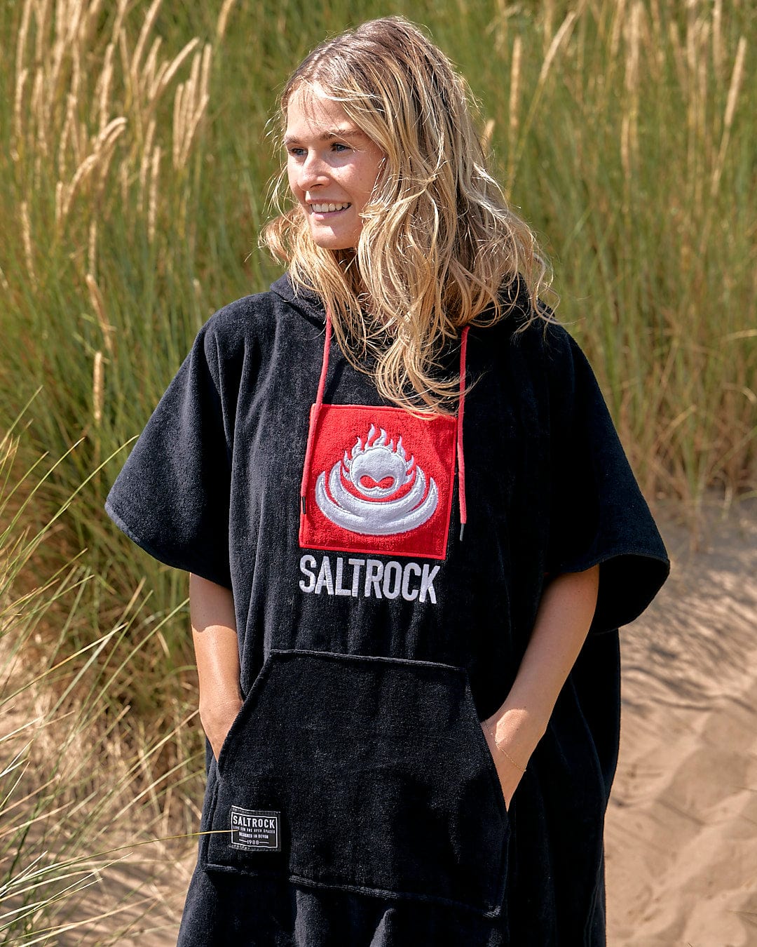 A person wearing a Saltrock Corp Changing Towel - Black standing on a sandy beach with tall grass in the background.