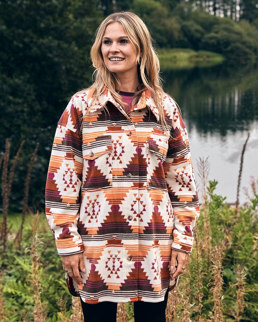 A woman wearing an Aztec print jacket, also known as the Dallyn - Womens Aztec Fleece Shacket - Orange by Saltrock, standing in front of a lake.