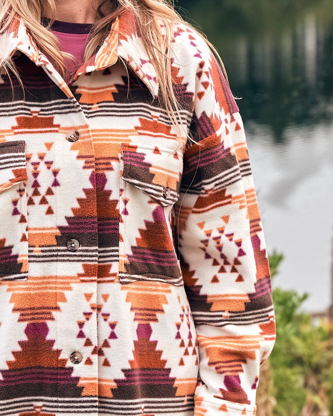 A woman wearing an Aztec print Saltrock Dallyn - Womens Aztec Fleece Shacket - Orange near a lake.