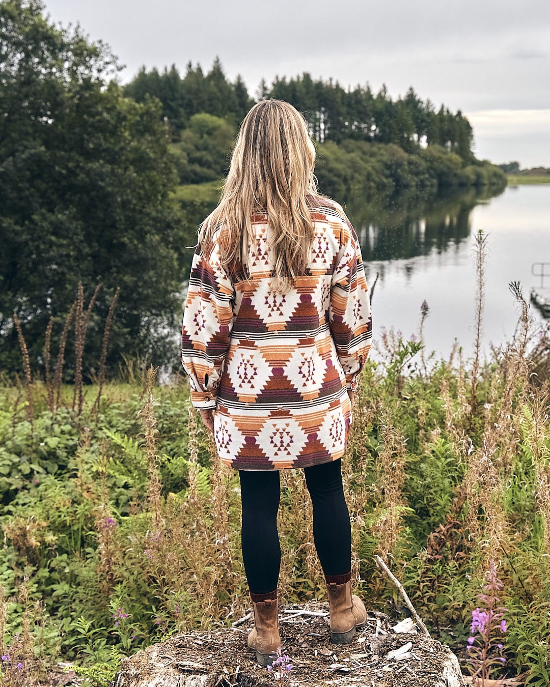A woman wearing the Saltrock Dallyn - Womens Aztec Fleece Shacket - Orange is standing on top of a rock, gazing at a lake.
