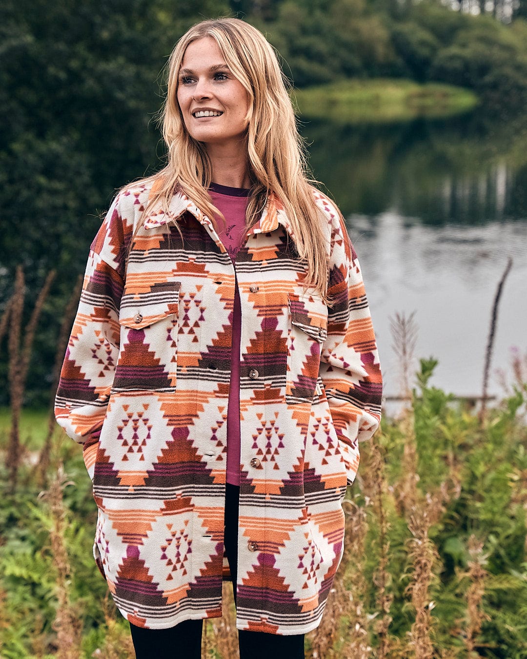 A woman wearing a Saltrock Dallyn - Womens Aztec Fleece Shacket - Orange, standing in front of a lake.