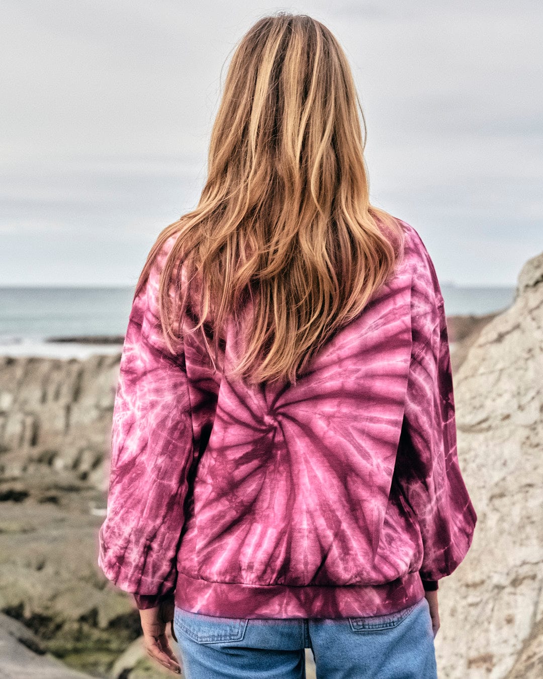 A fashionista wearing a Saltrock Dawn - Womens Tie Dye Sweatshirt - Pink looks at the ocean.