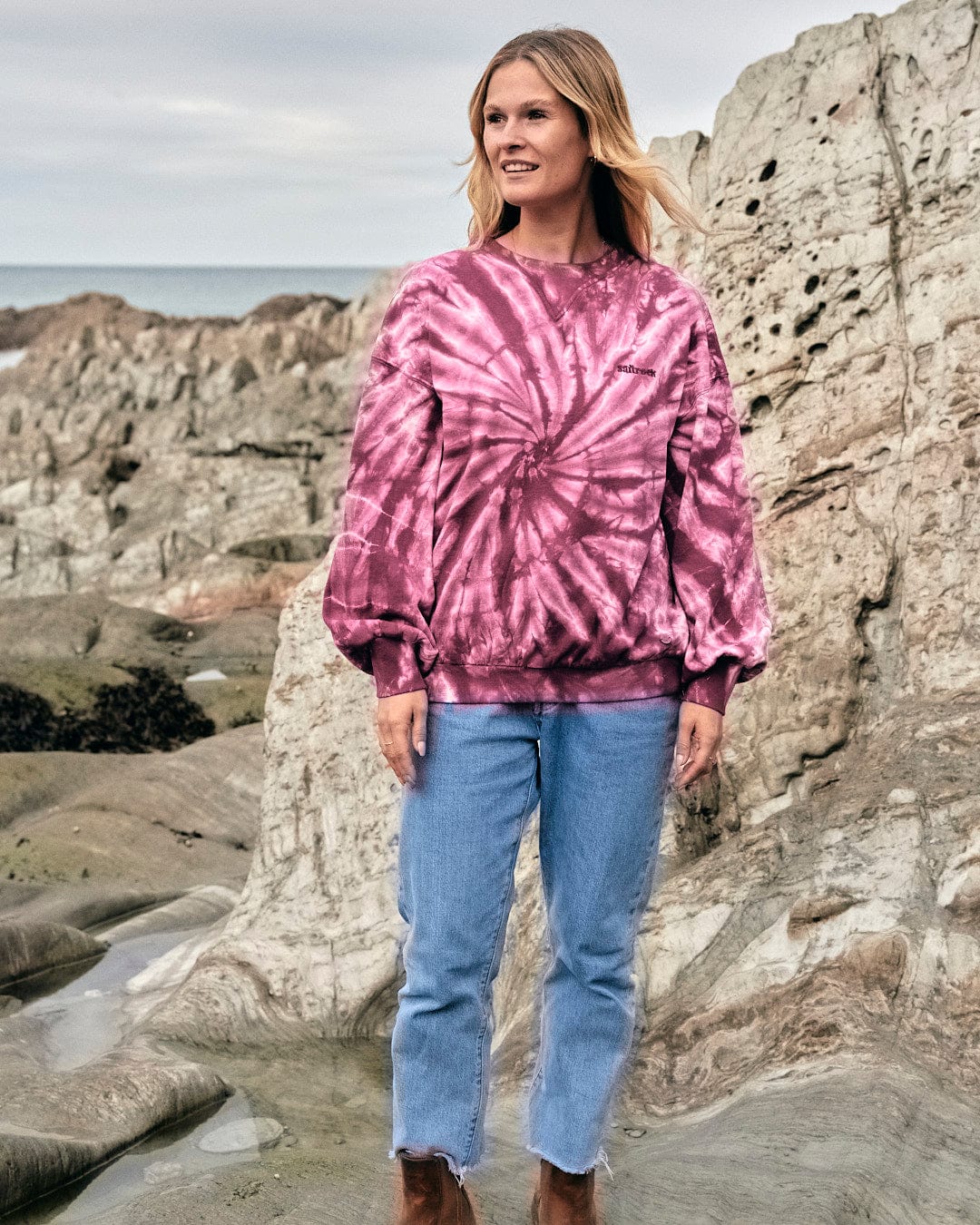 A fashionista wearing a pink tie dye sweatshirt featuring Saltrock branding, standing on rocks.