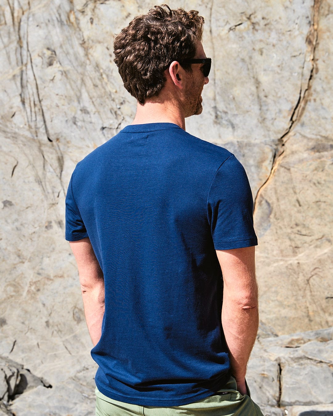 A man wearing a Saltrock Diamond Scene - Mens Short Sleeve T-Shirt - Blue is standing in front of a rock.