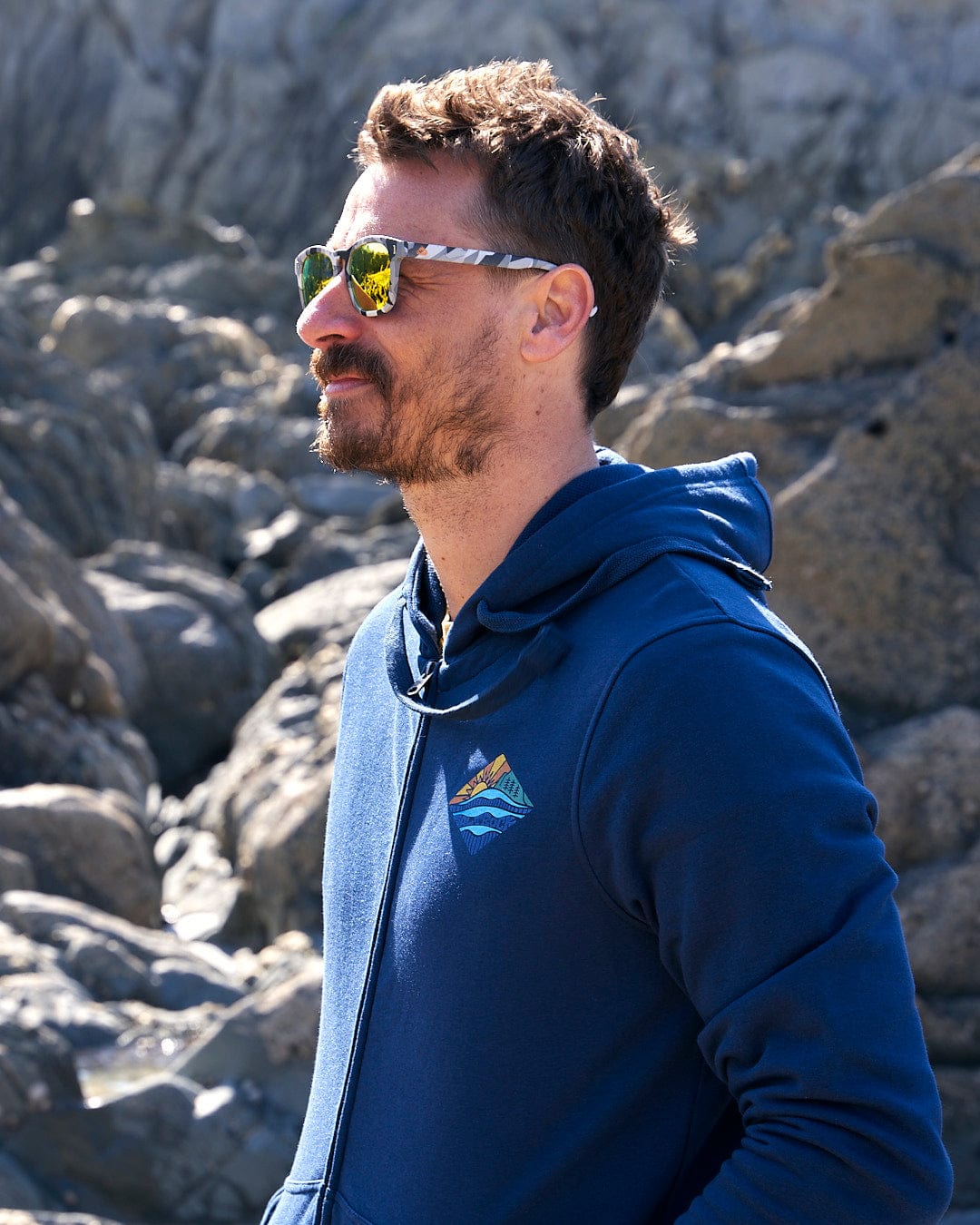A man wearing the Saltrock Diamond Scene - Mens Zip Hoodie - Blue and sunglasses standing next to rocks.