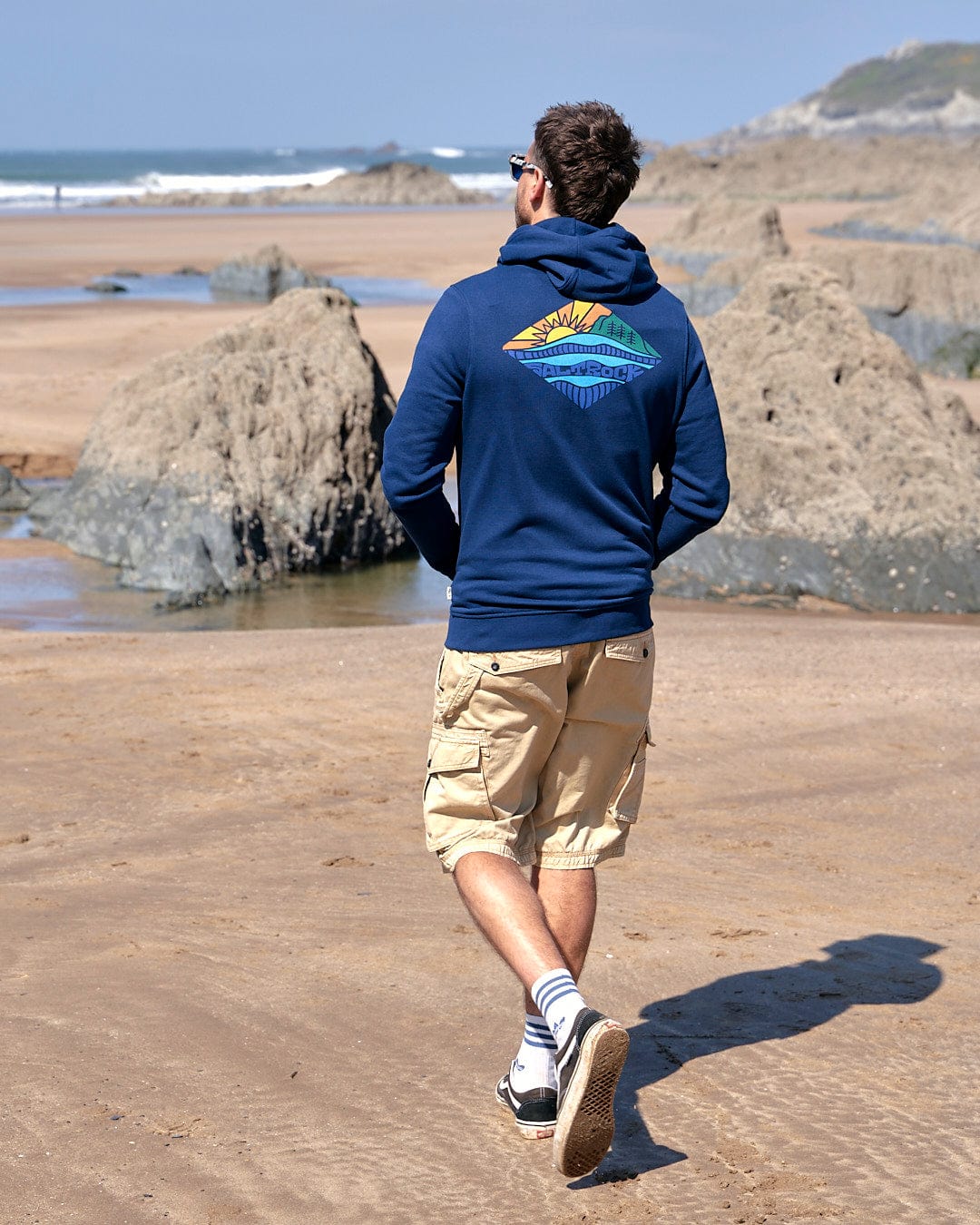 A man walking on the beach wearing a Saltrock Diamond Scene - Mens Zip Hoodie in Blue.