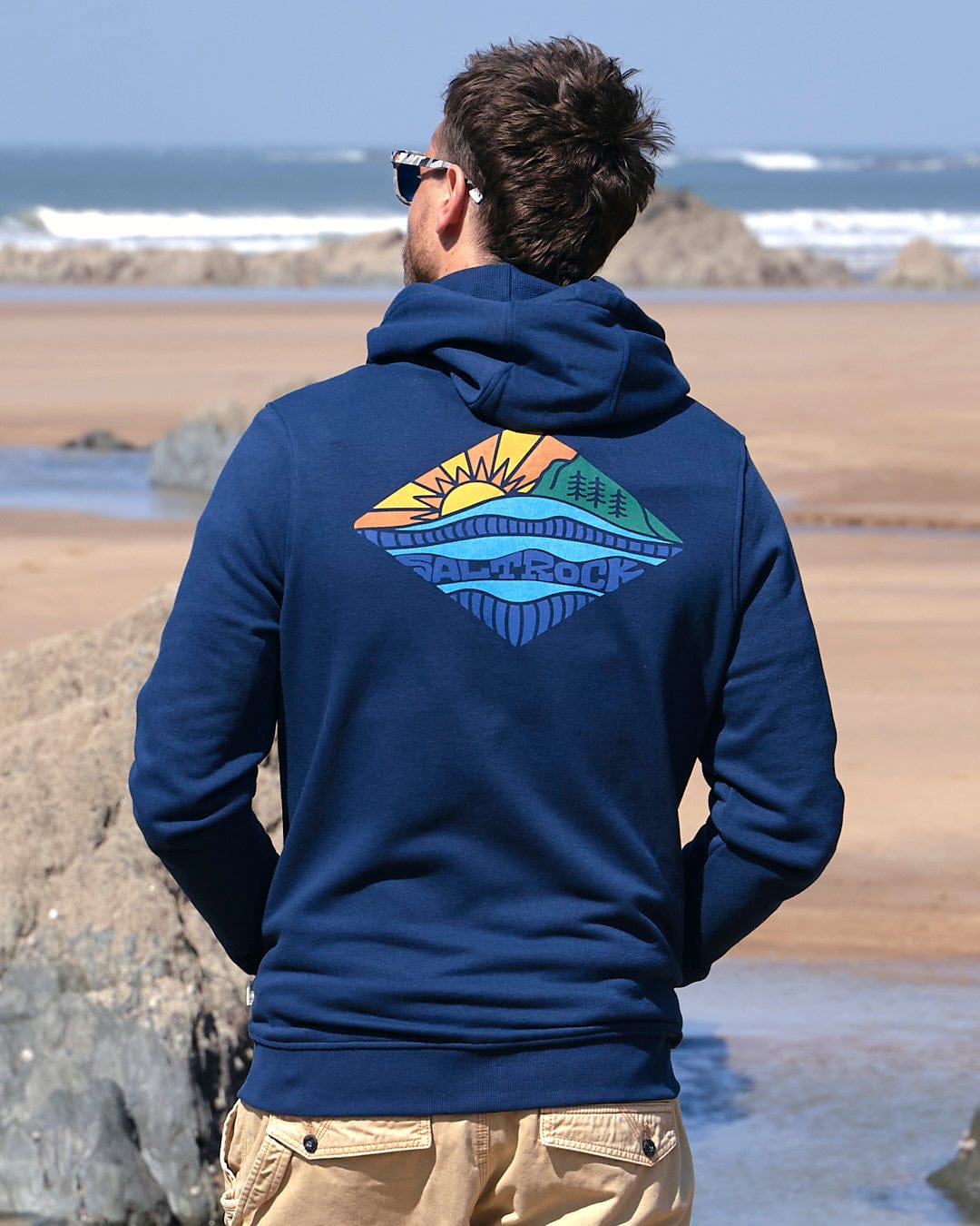 A man is standing on the beach wearing a Saltrock Diamond Scene - Mens Zip Hoodie - Blue.