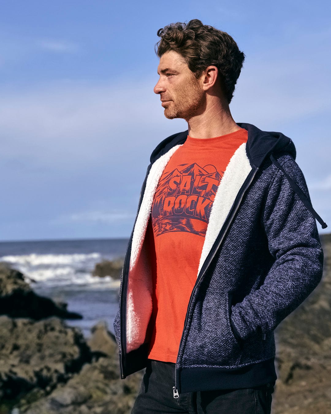 A Saltrock Dillan - Mens Lined Hoodie - Dark Blue-wearing man, with his hands tucked into the front pockets, standing on rocks near the ocean.