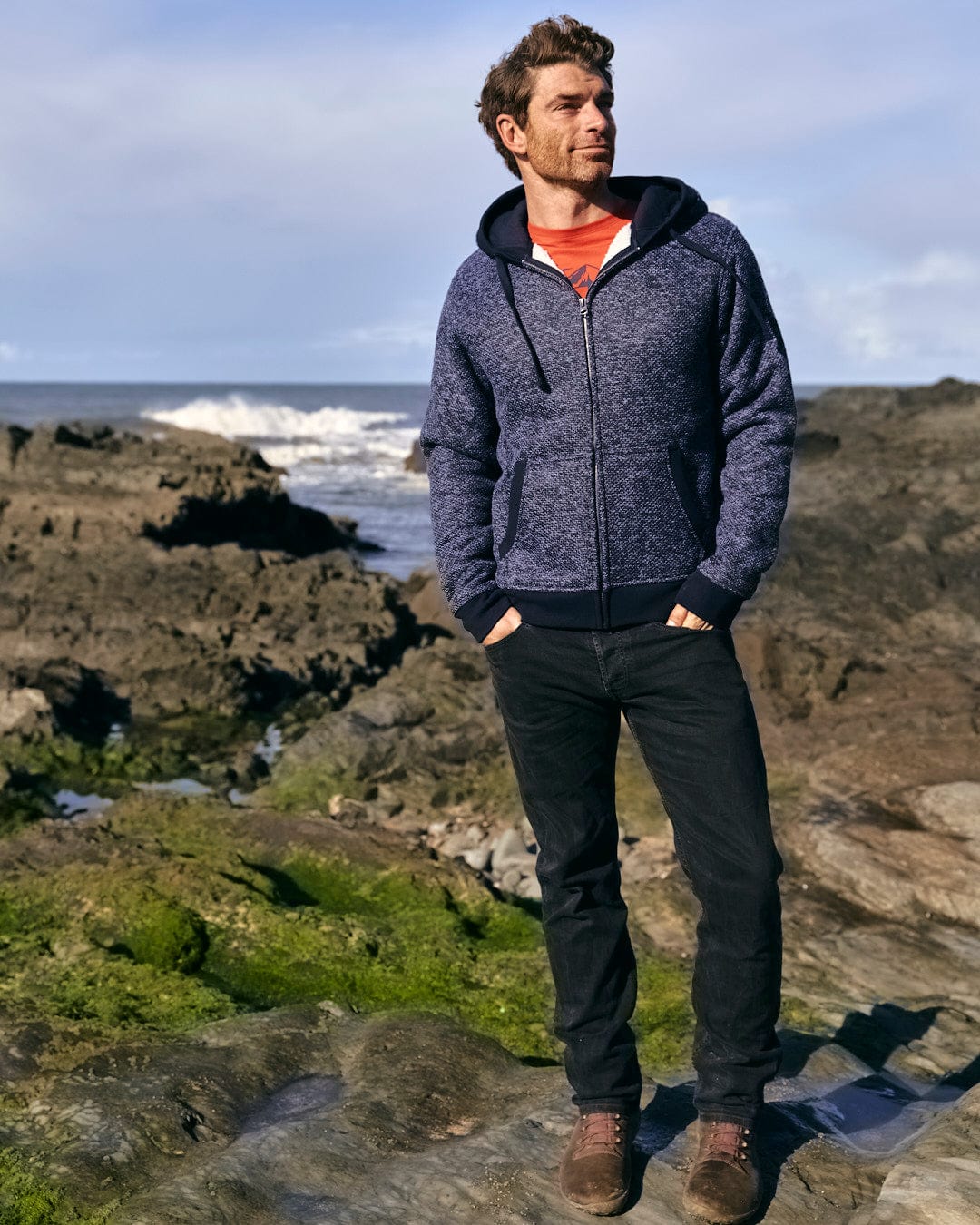 A man wearing a Saltrock Dillan - Mens Lined Hoodie - Dark Blue stands on rocks next to the ocean, with his hood up.
