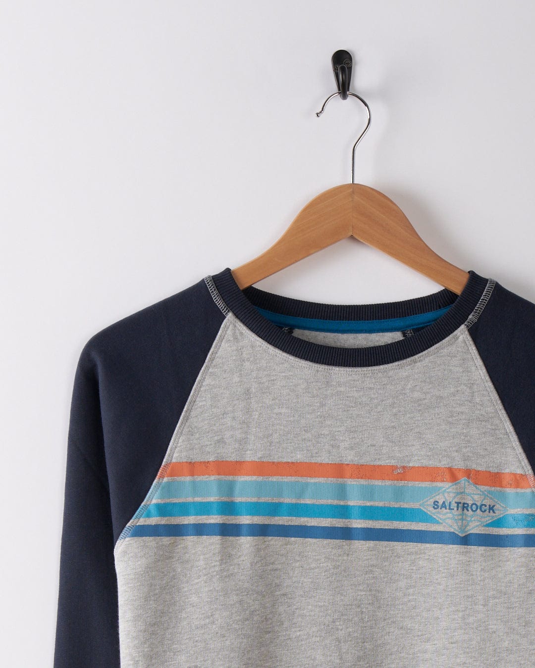 The Saltrock Dimensions Stripe Men's Sweat in blue/grey features orange and blue stripes, a crew neck, and raglan sleeves, displayed on a wooden hanger against a white wall.