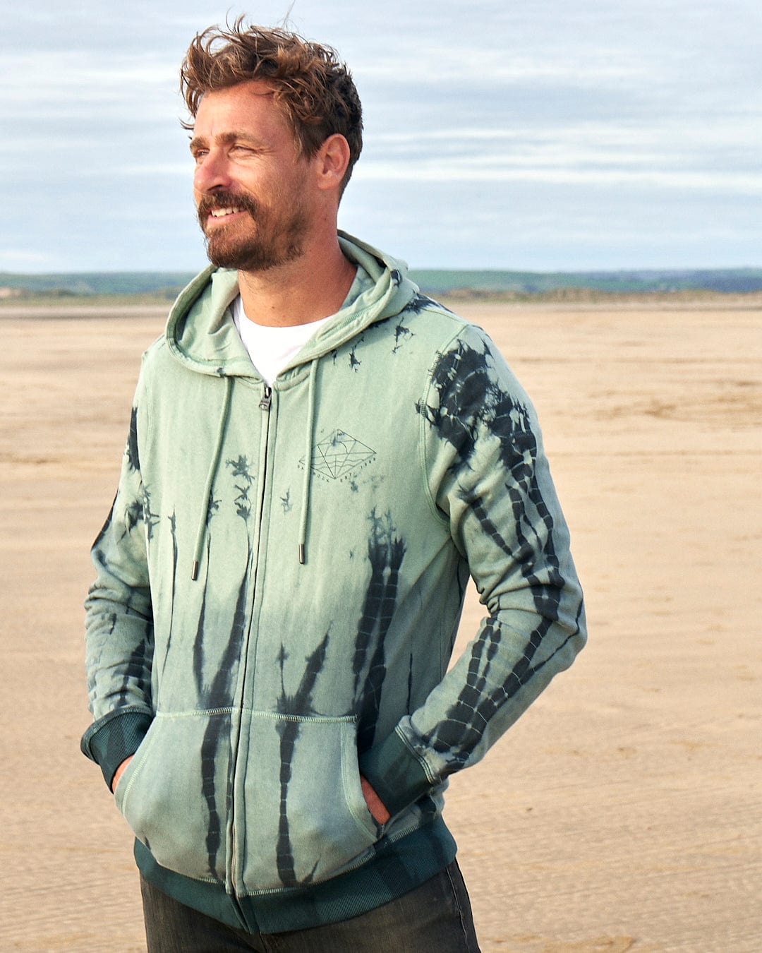 A man on the beach wearing a Saltrock Dimensions - Mens Zip Tie Dye Hoodie - Green.