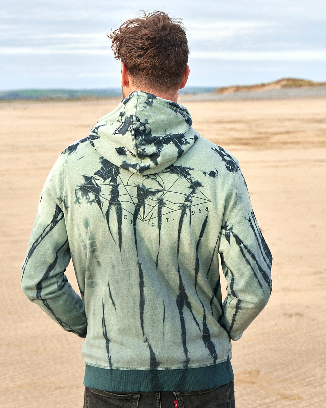 A man standing on the beach wearing a Saltrock - Dimensions Mens Zip Tie Dye Hoodie - Green.