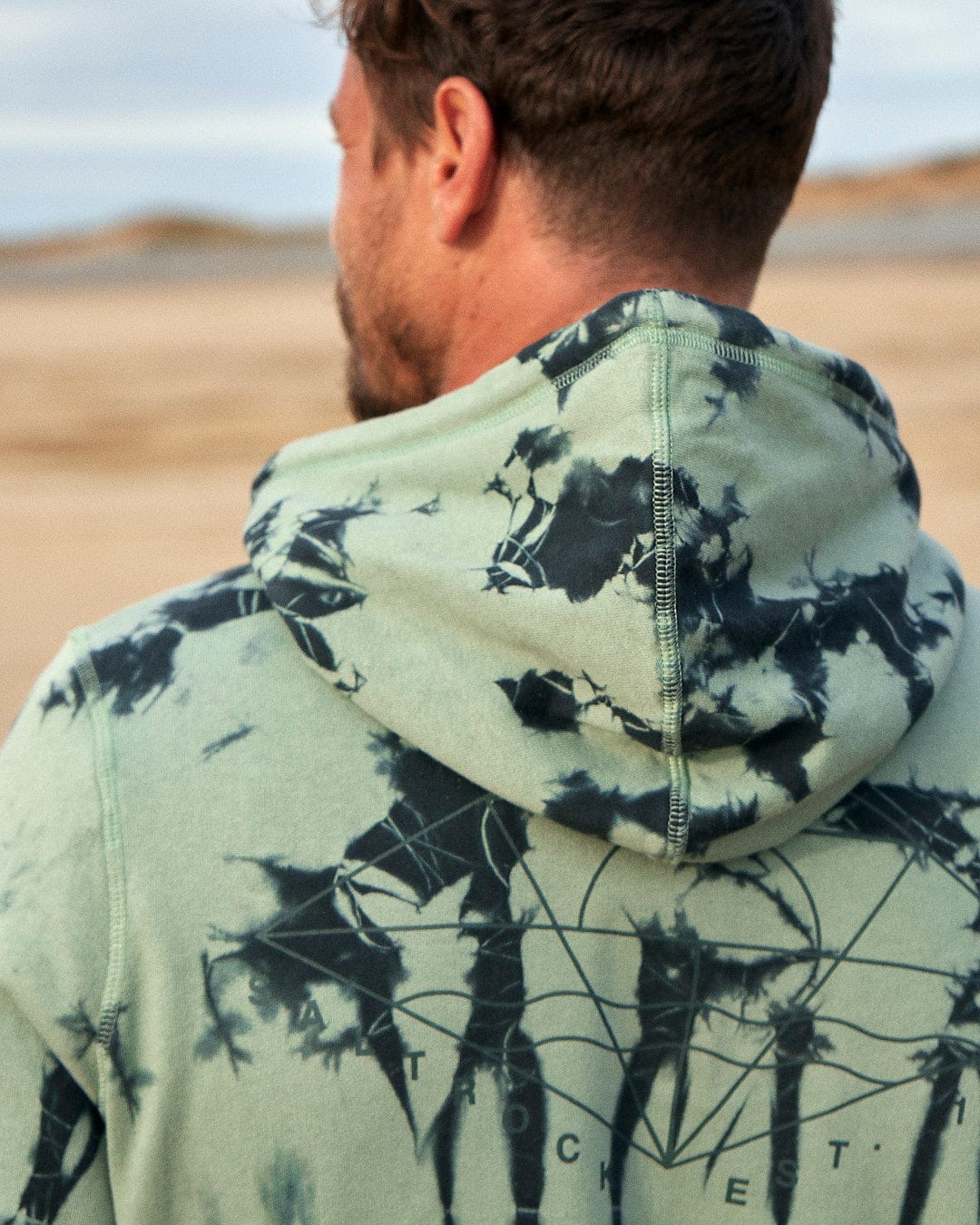 A man is wearing a Saltrock Dimensions - Mens Zip Tie Dye Hoodie - Green on the beach.