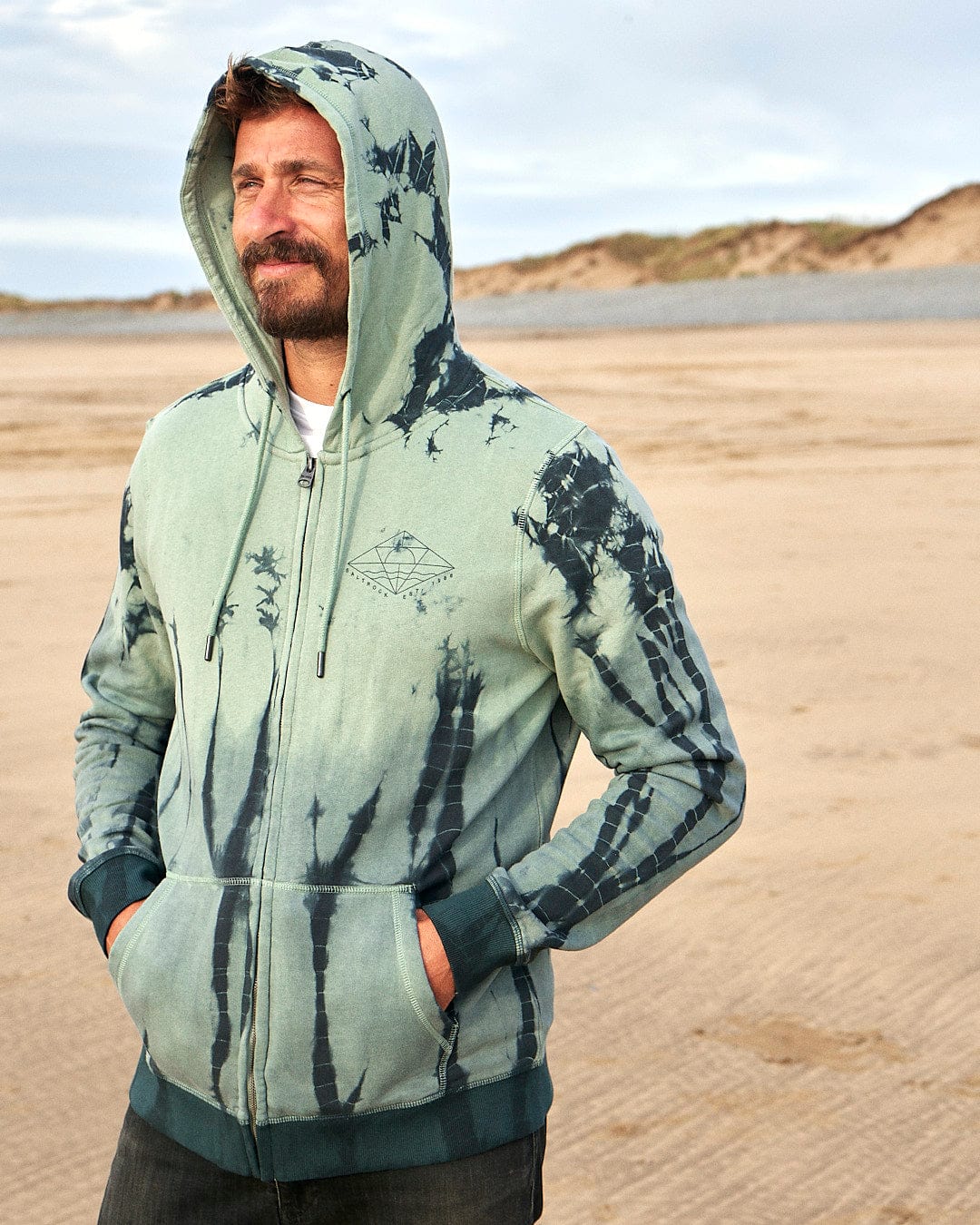 A man wearing a Saltrock Dimensions - Mens Zip Tie Dye Hoodie - Green standing on a beach.