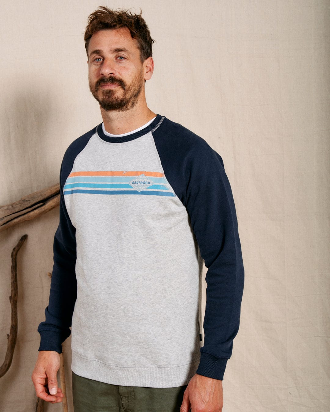 A man stands indoors wearing the Saltrock Dimensions Stripe Men's Sweat in blue/grey, featuring multicolored chest stripes and a subtle logo, paired with olive green pants.