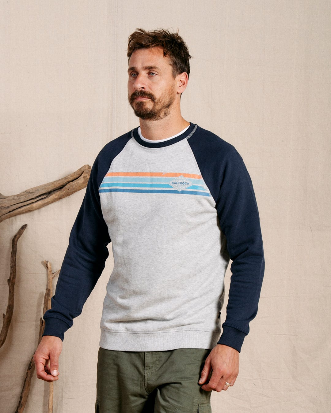 A man with short brown hair and a beard wears the Saltrock Dimensions Stripe Men's Sweat in blue/grey with colorful chest stripes and green pants, standing indoors near driftwood.