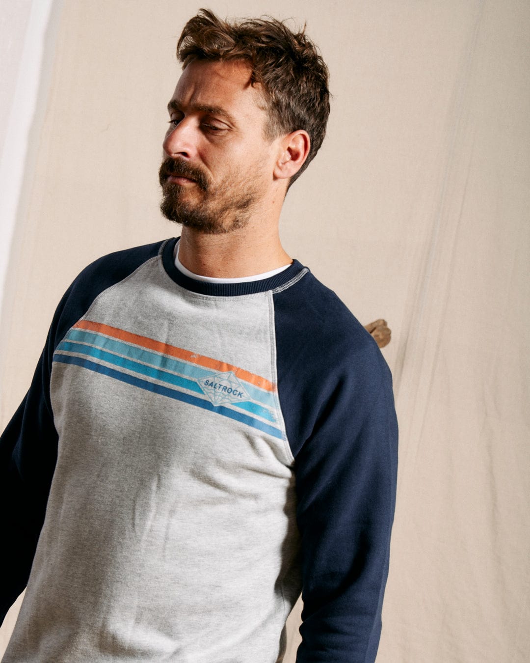 A man with short brown hair and a beard wears the Saltrock Dimensions Stripe Men's Sweat in blue/grey, standing before a beige backdrop.