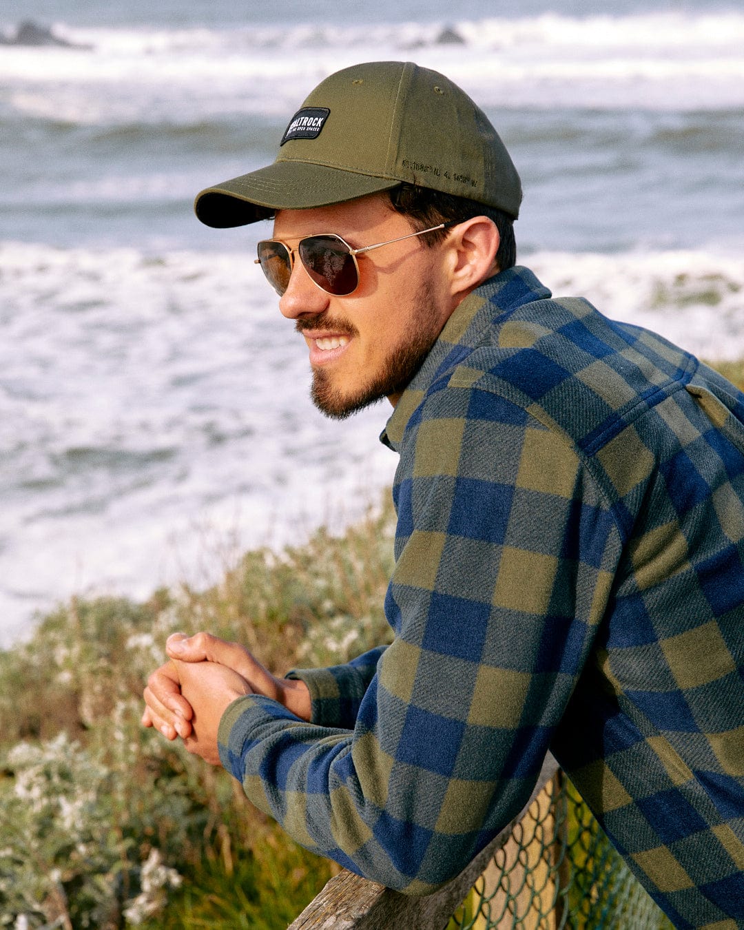 Man in sunglasses and a Saltrock Dockyard - Cap - Green embroidered plaid jacket leaning on a fence by the sea, looking away thoughtfully.