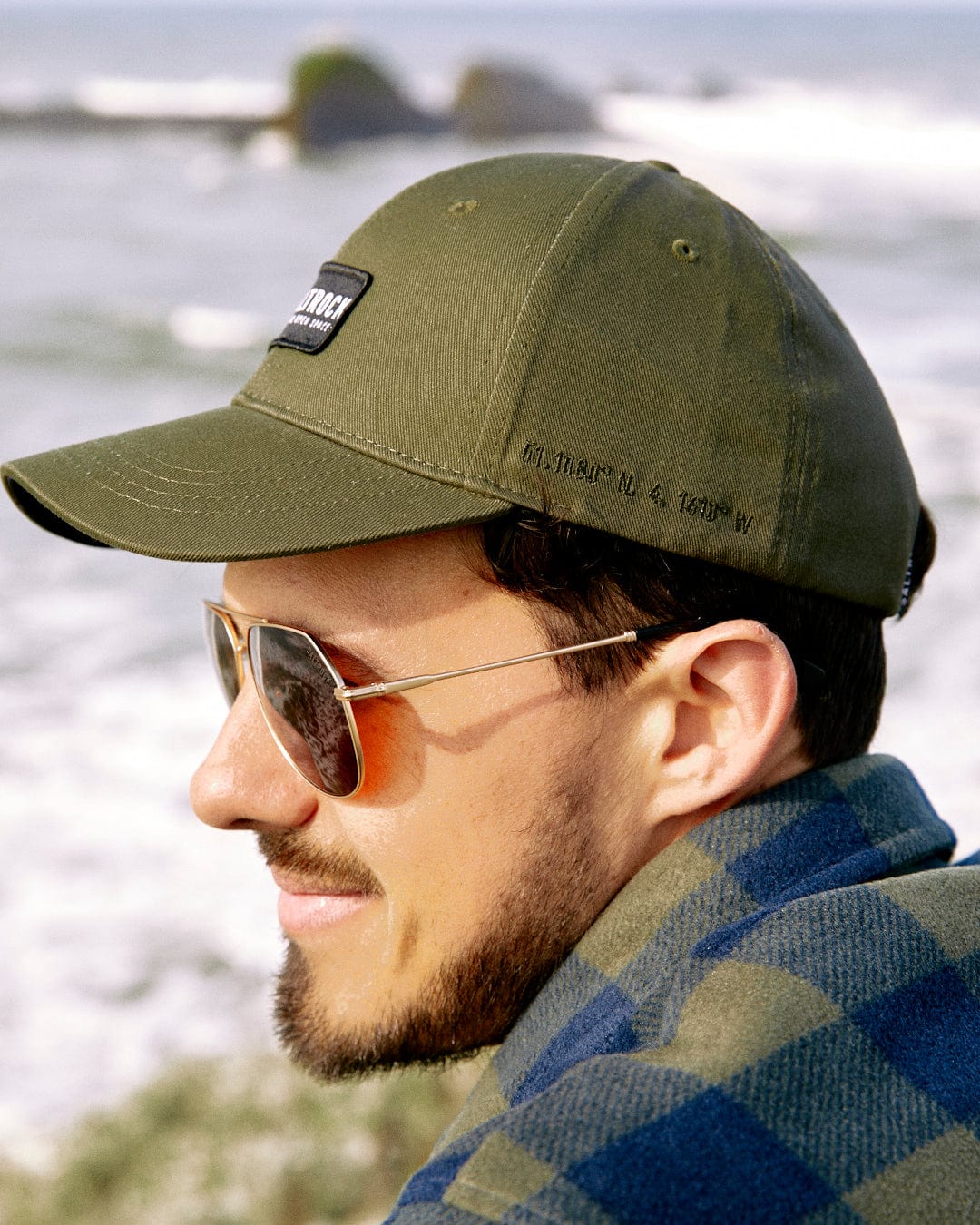 Profile view of a young man wearing a Saltrock Dockyard - Cap - Green with an adjustable back and sunglasses, with a blurred natural background.