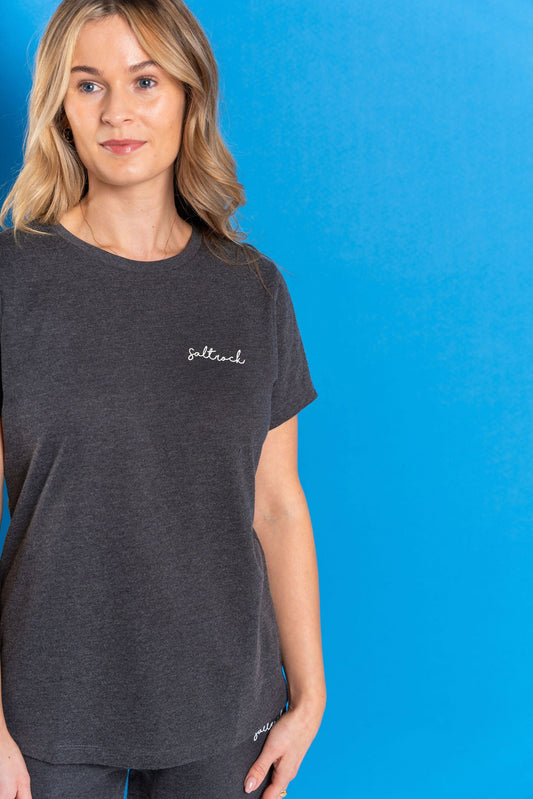 A woman stands before a blue background in the Saltrock Velator Women's Short Sleeve T-Shirt in dark grey, featuring Saltrock branding on the chest and crafted from recycled materials for a soft, peached feel.