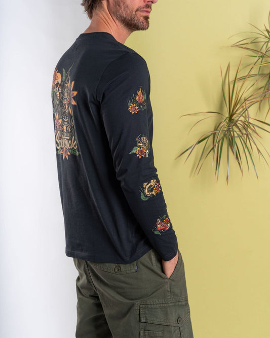 A man in a Saltrock Lantern dark grey long-sleeve t-shirt with colorful floral designs, made of 100% cotton, stands sideways with his hand in his pocket next to a plant against a yellow wall.