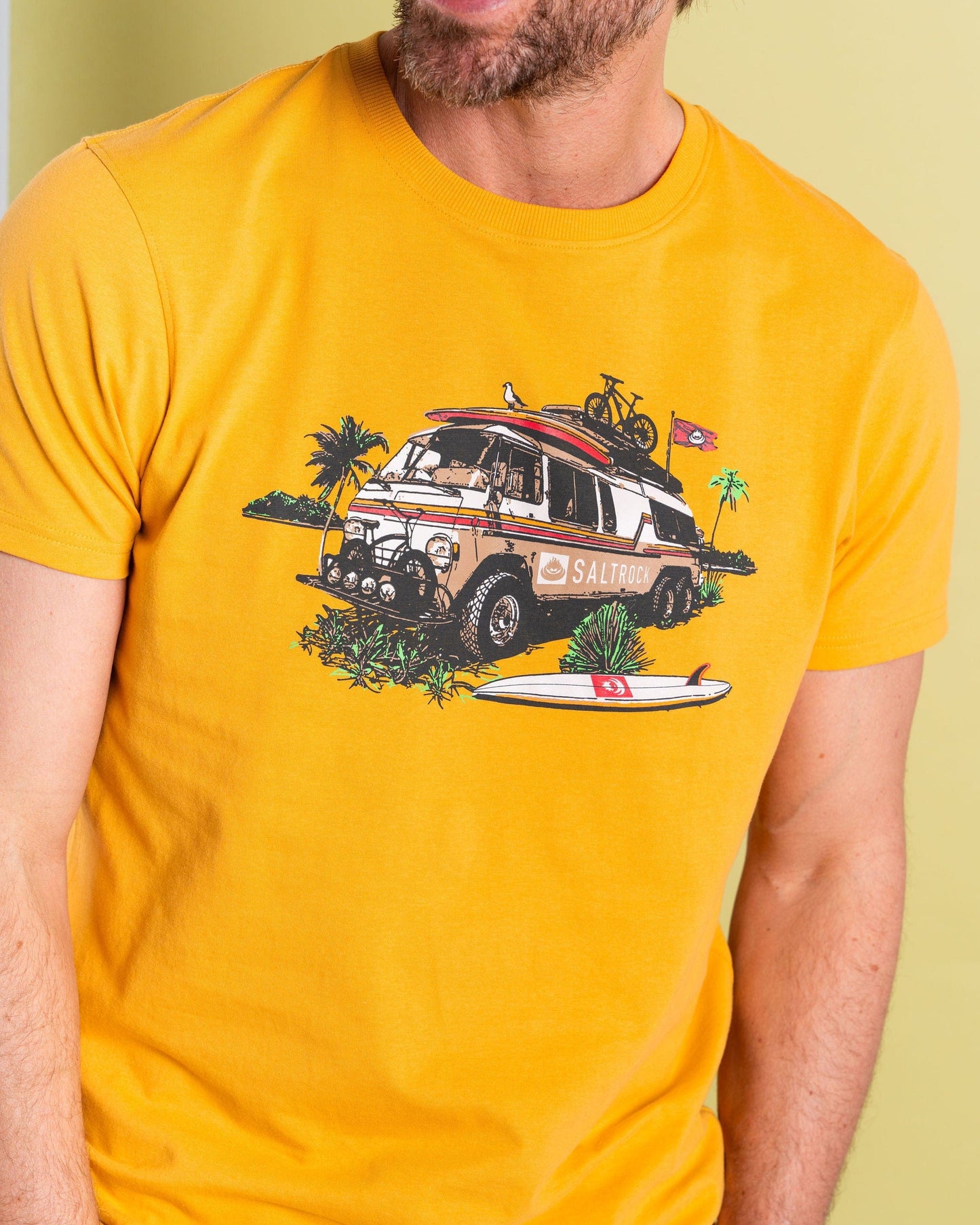 A man wearing a Saltrock "Retro Camper" yellow t-shirt featuring a graphic of a camper, palm trees, bicycle, surfboard, and flags.