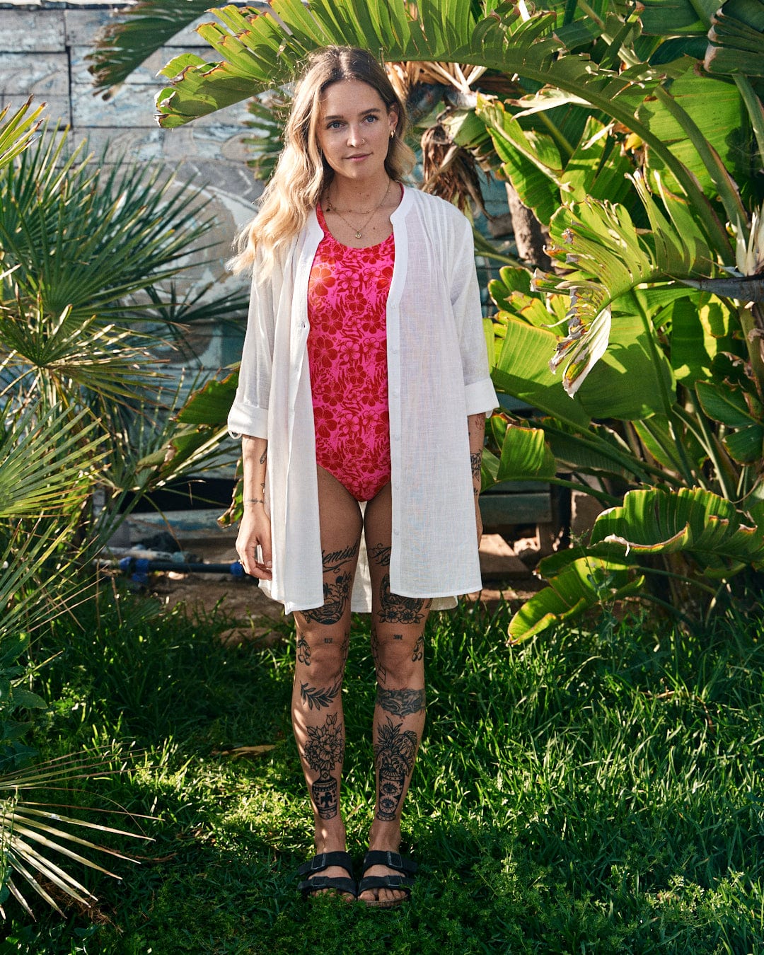 Woman in a pink swimsuit and Saltrock lightweight cotton shirt standing in front of lush green foliage, showing off detailed leg tattoos.