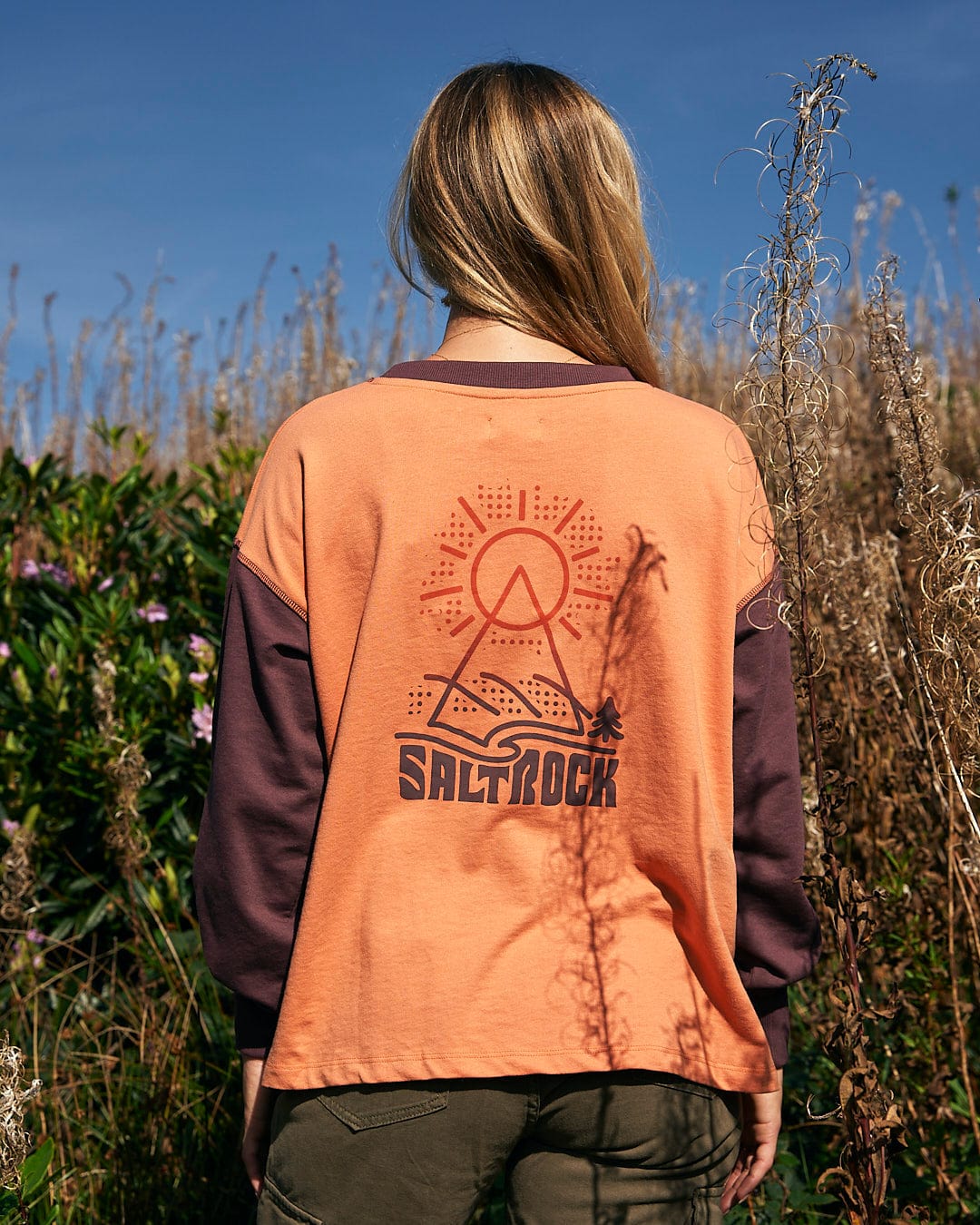 A woman with a comfortable style standing in a field wearing a Saltrock Elissa - Womens Boxy Sweat - Orange t-shirt.