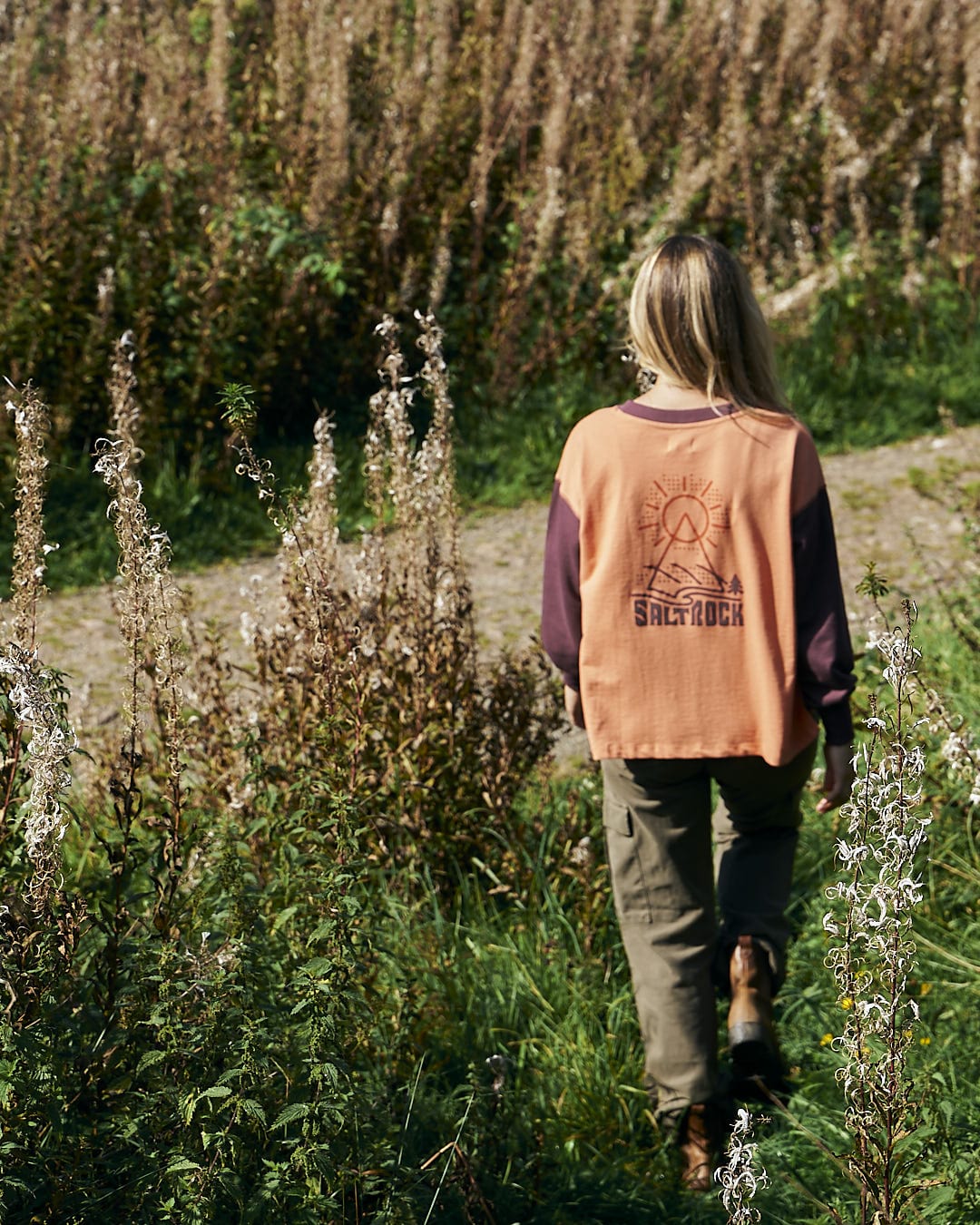 A stylish woman walking through a field of tall grasses wearing the Elissa - Womens Boxy Sweat - Orange by Saltrock.