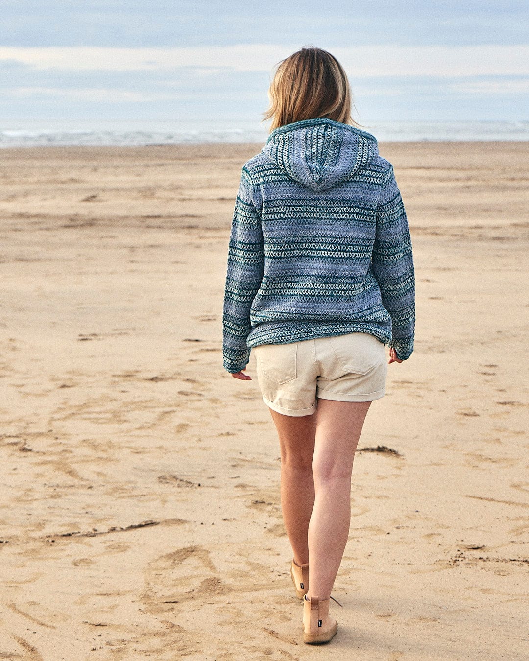A woman walking on the beach wearing a Saltrock Erin - Womens Hooded Knit in Light Green hoodie and shorts.