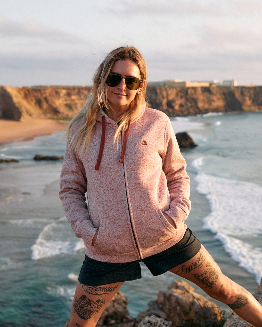 A woman in a Farley - Womens Borg Lined Hoodie in Pink from Saltrock is standing on a cliff, gazing at the ocean.