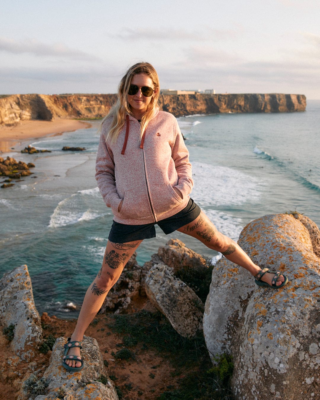 A woman in a Farley - Womens Borg Lined Hoodie in Pink by Saltrock is posing on rocks by the ocean.