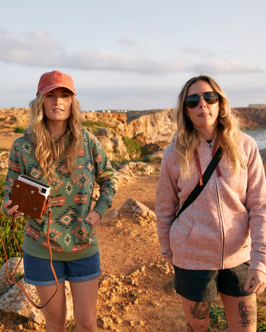 Two women standing on a rocky path near the ocean, wearing Saltrock pink Farley - Womens Borg Lined Hoodies with drawstring hoods.