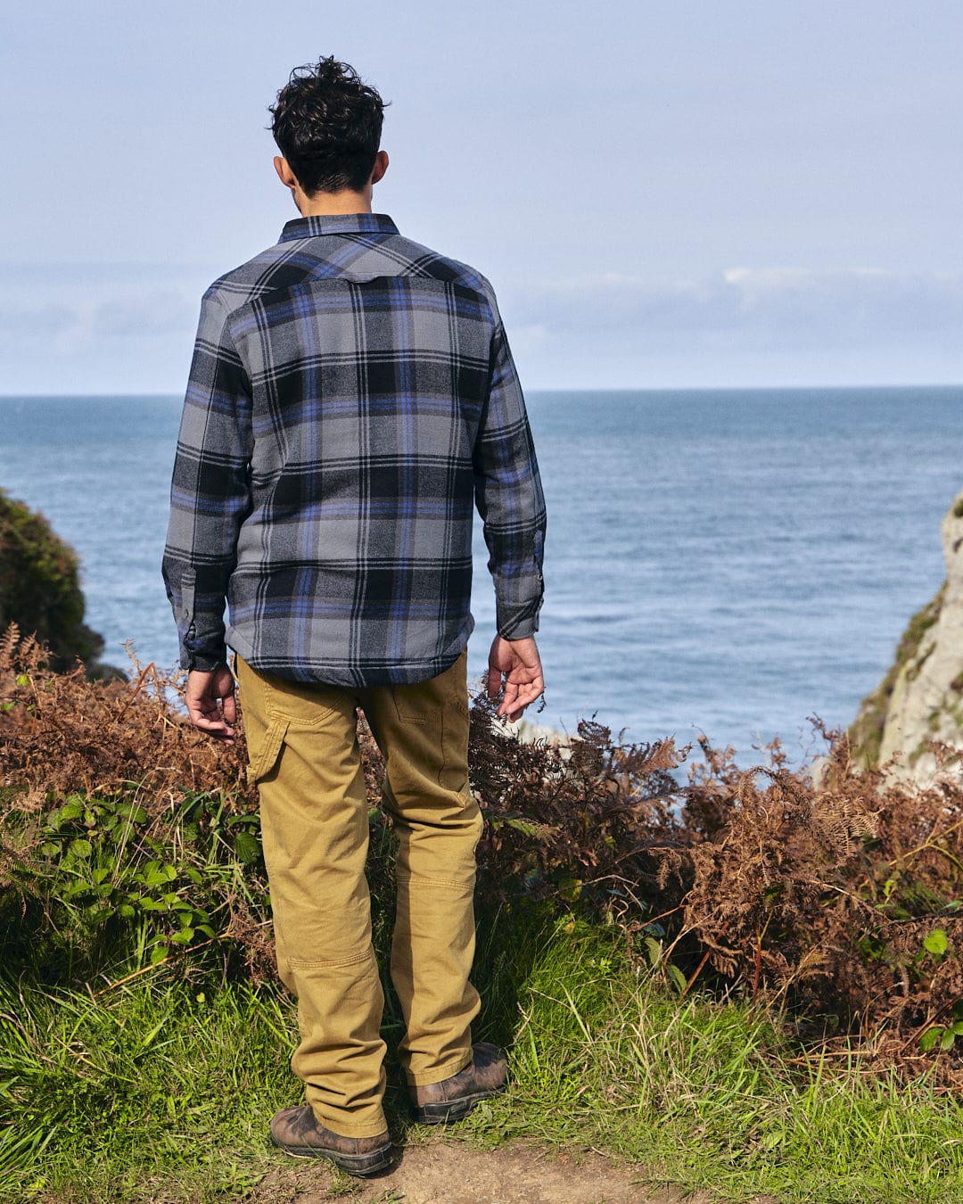 A stylish man in Saltrock's Woody - Mens Sherpa Lined Shacket - Grey attire is standing on a path, gazing at the warm expanse of the ocean.