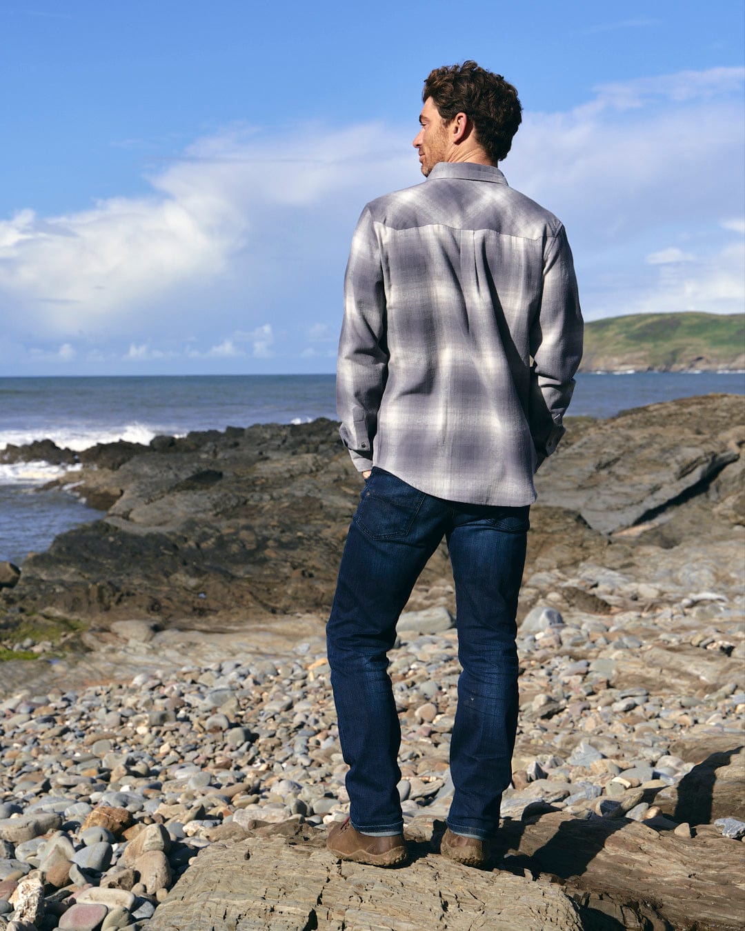 A man standing on rocks by the ocean, wearing a Farris - Mens Long Sleeve Check Shirt - Grey made by Saltrock for maximum comfort.