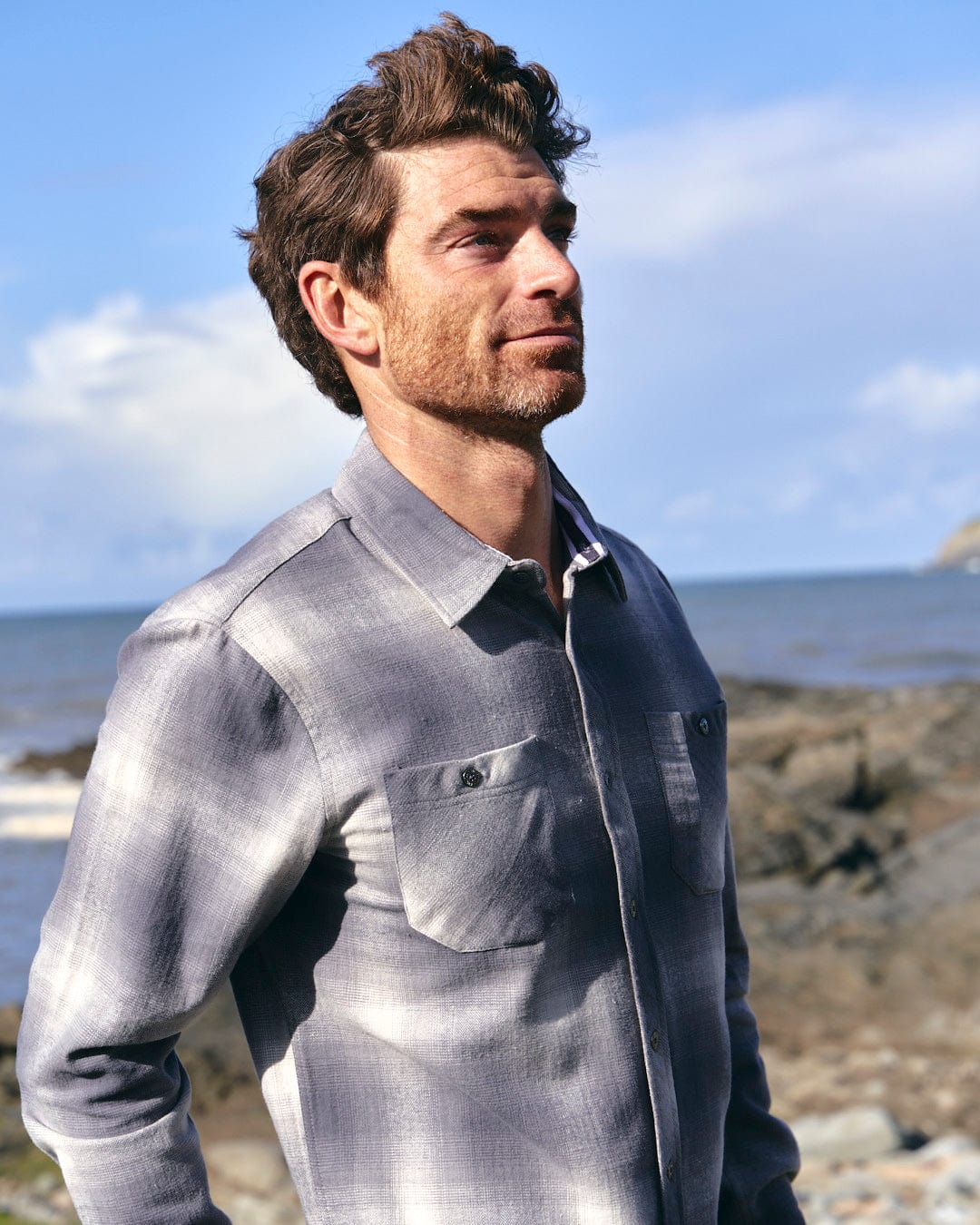 A man standing on a rocky beach wearing a Farris - Mens Long Sleeve Check Shirt - Grey with chest pockets, made by Saltrock.