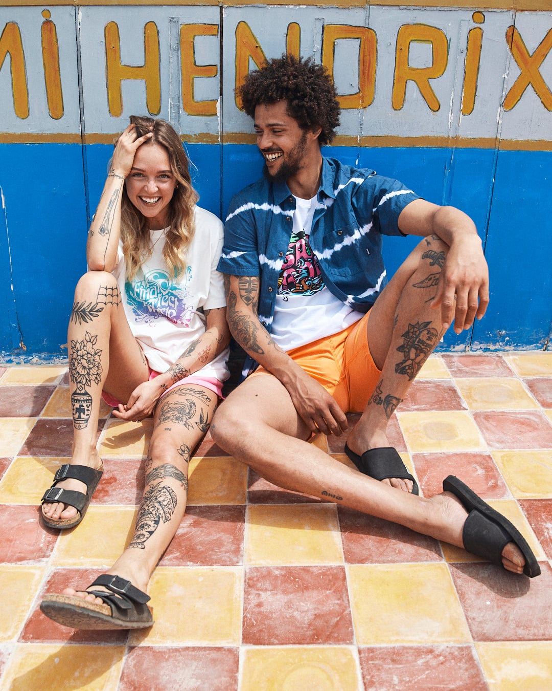 A man and woman with tattoos sitting happily together in front of a colorful wall with the multi-colored slogan "hendrix" painted on it wearing Saltrock's Find The Perfect Wave - Womens Short Sleeve T-Shirt.