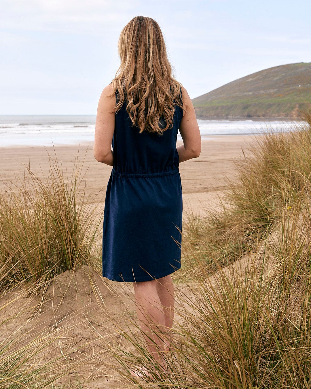 A woman standing on the beach in a Saltrock Flax - Womens Tie Vest Dress - Blue.