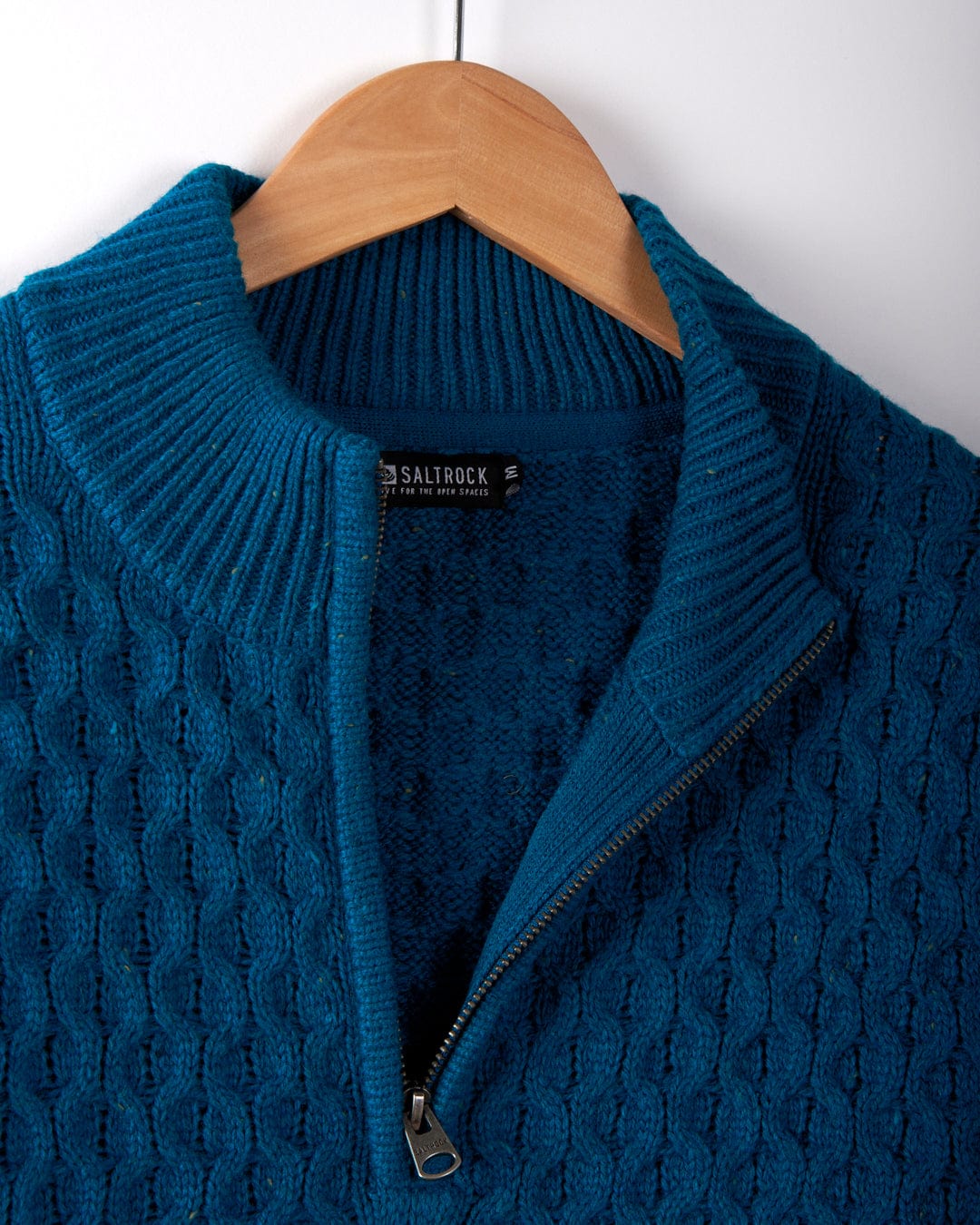 Close-up of the Saltrock Flint Men's 1/4 neck zip knit sweater in blue, featuring a textured cable knit pattern, ribbed collar, and partially unzipped front, hanging on a wooden hanger.