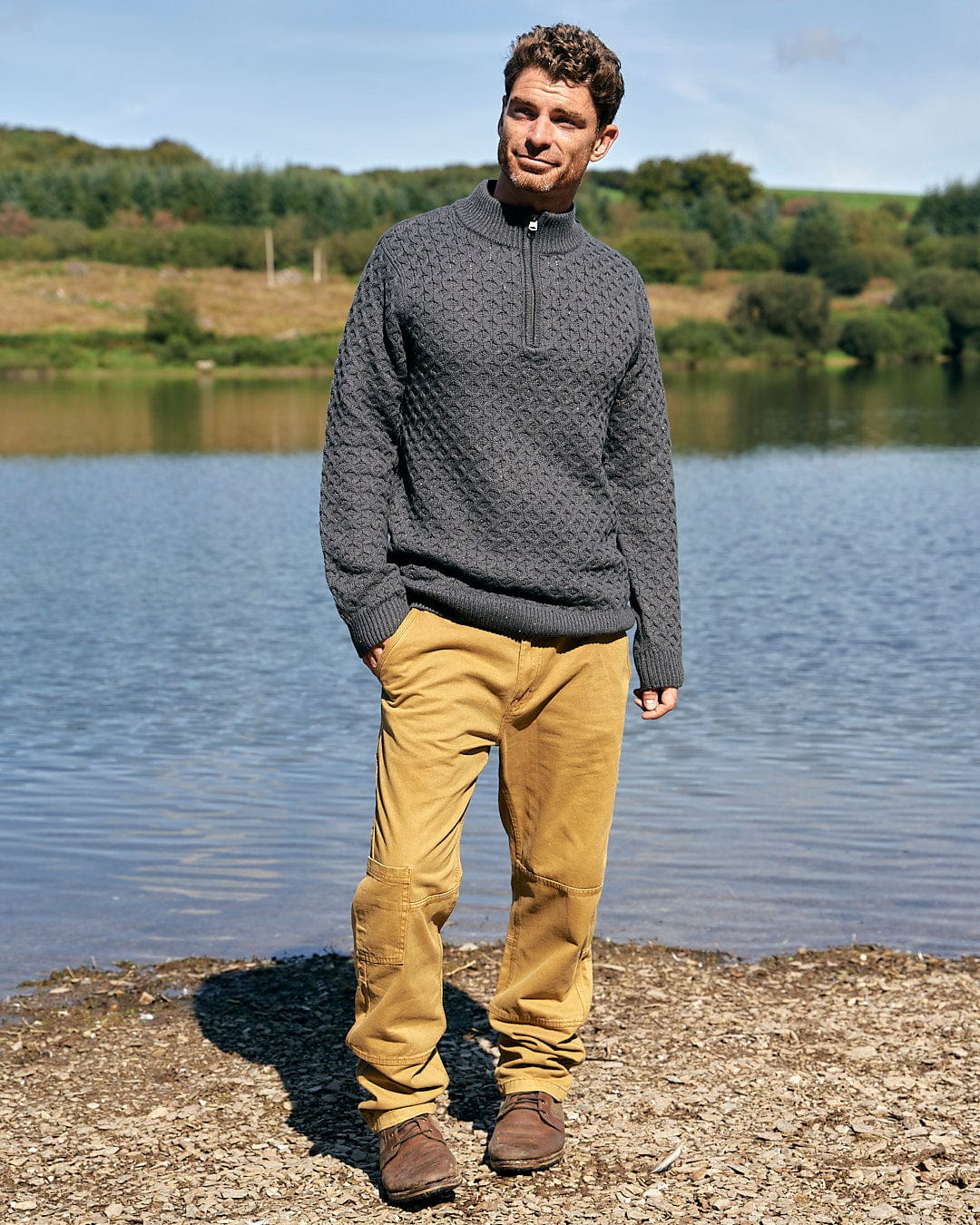 A man wearing a Flint - Mens 1/4 Neck Zip Knit - Black sweater by Saltrock and khakis, standing next to a lake.