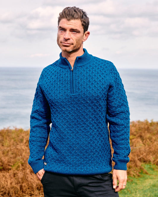 A man stands by the coast wearing Saltrock's Flint - Mens 1/4 Neck Zip Knit in blue and black pants, with brown vegetation and the ocean behind him.