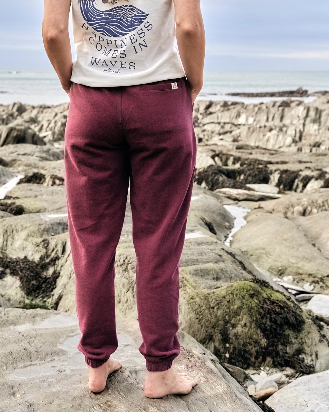 A stylish woman wearing Saltrock maroon joggers and a t-shirt standing on rocks near the ocean.