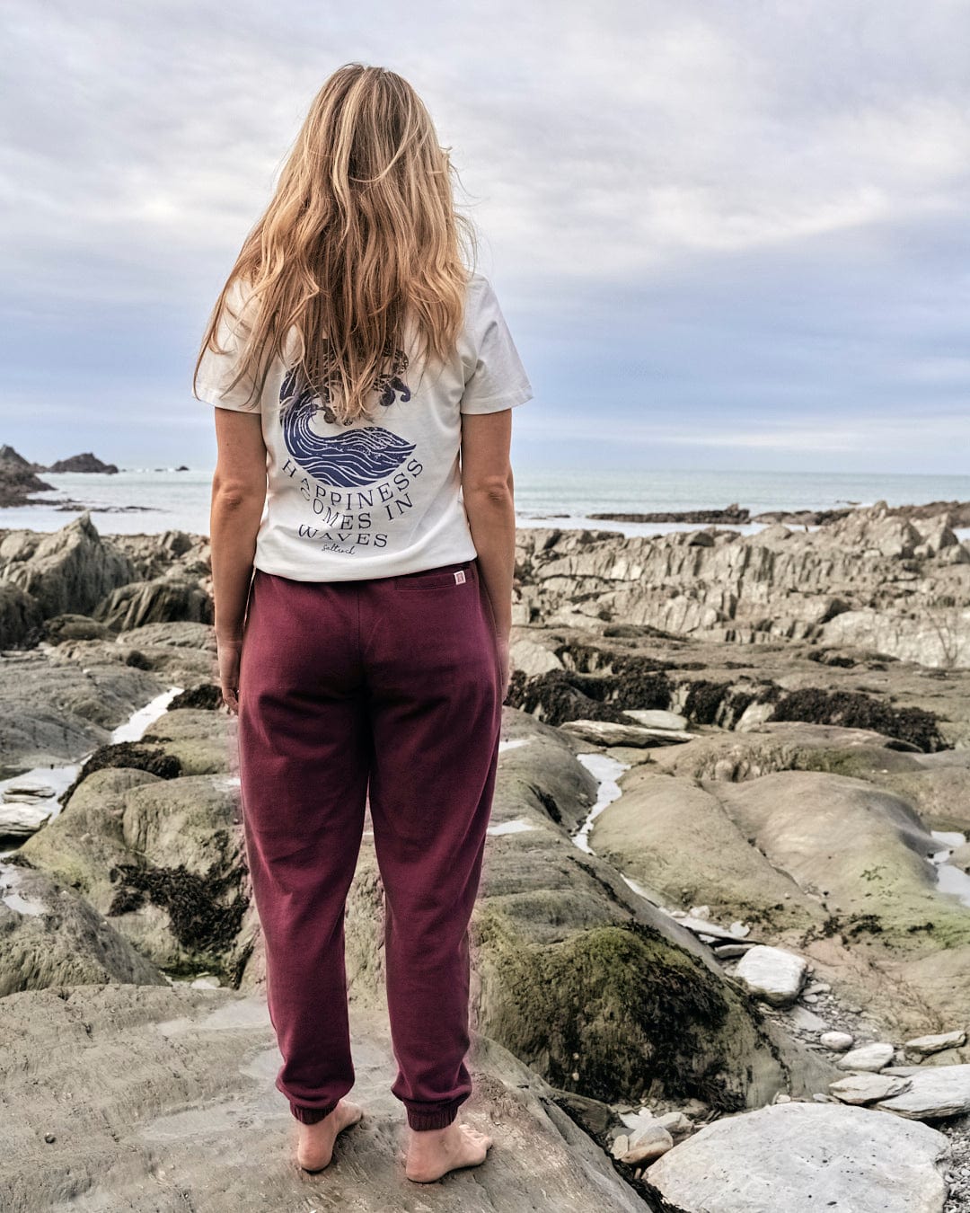 A stylish woman wearing Saltrock Flo - Womens Jogger - Purple sweatpants is standing on rocks near the ocean.