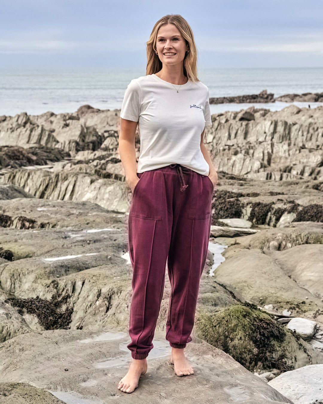 A woman wearing Saltrock's Flo - Womens Jogger - Purple standing on a rocky beach.