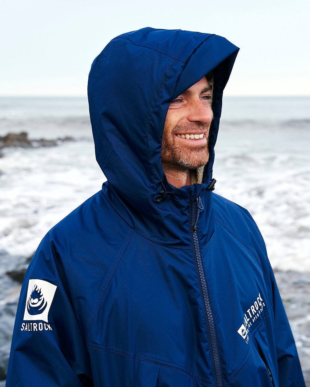 A man wearing a blue hooded jacket stands near the ocean, smiling. His jacket, a Changing Robe - Blue with the Saltrock logo on the sleeve and chest, looks perfect for any weather. Waves are visible in the background.