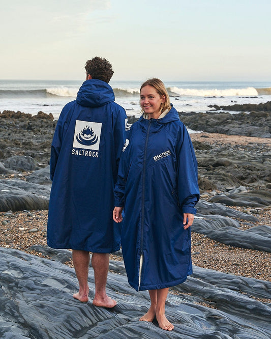 Two people wearing Saltrock Recycled Four Seasons Changing Robes in Blue stand on a rocky beach, looking at each other with the ocean and waves in the background.