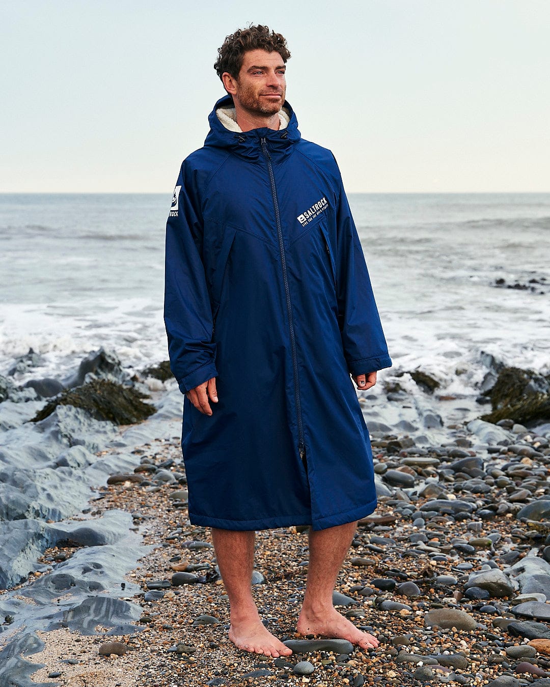 Man standing on a rocky beach wearing a Saltrock Recycled Four Seasons Changing Robe in Blue, with the ocean in the background.