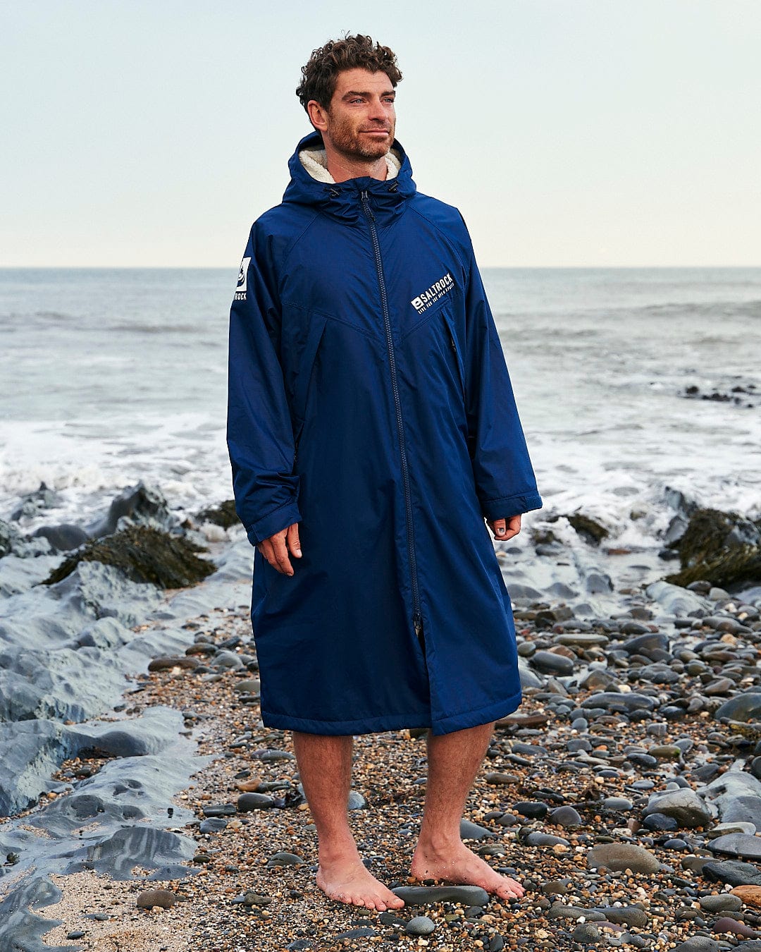 A person stands barefoot on a rocky beach, wearing a long blue Saltrock Changing Robe - Blue with a hood. The ocean waves are crashing in the background.